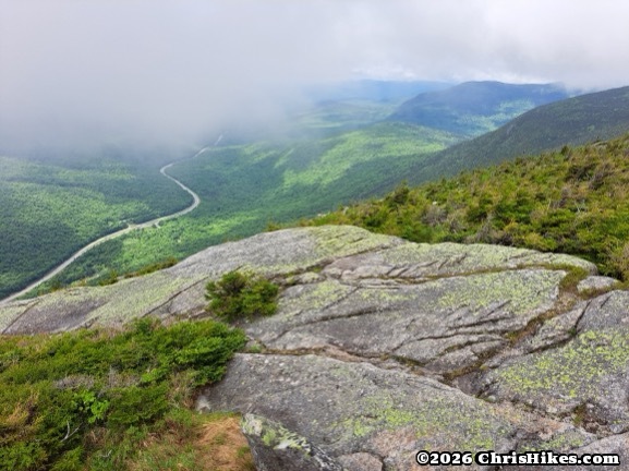 photo of rocky cliff with cloud covered mountains in the background and a highway running through the valley