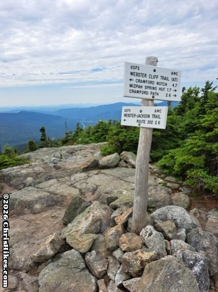 Two white rectangular signs with black letters mounted on a white post; windswept trees and mountains in the background