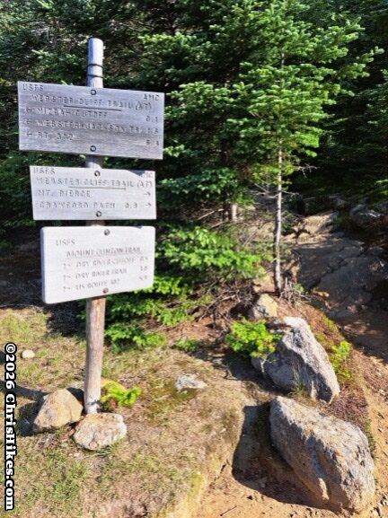 Three weather worn signs on a post, evergreen trees and hiking trail in background