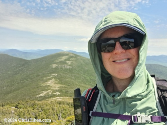 Photograph of person wearing hoodie, hat, and sunglasses with mountain summit in background