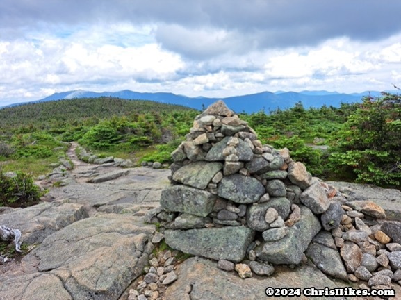 photograph of pile of rocks on top of a mountain with trees and mountains in the distance